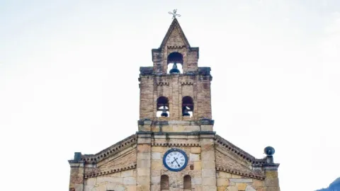  Primer plano de la parte alta de la iglesia en piedra en la que sobresale la torre con el campanario y el reloj.