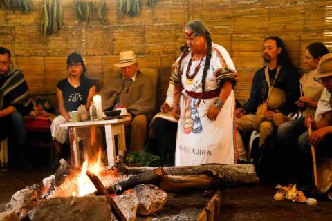 Una escena en el interior de una estructura rústica con paredes de bambú. Un grupo de personas se encuentran sentadas a lo largo de las paredes. Destaca una figura femenina de pie, vestida con un atuendo tradicional blanco adornado con símbolos y la palabra "Guajira". Lleva el pelo en trenzas y luce un tocado y collares coloridos. Delante de la figura, hay una fogata encendida con leños gruesos y rocas alrededor, que ilumina la escena con llamas y luz cálida. 