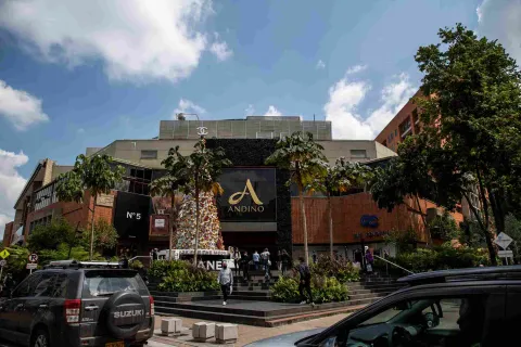 Facade of a building with at least three stories, made of brick and concrete, featuring a large, gold letter A and the word ""Andino."" In front, there are stairs with people and a few cars nearby.