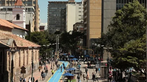 Un animado tramo peatonal de la Bogotá, donde la tradición y la modernidad conviven en un mismo encuadre. A la izquierda se aprecia la arquitectura colonial en ladrillo y teja de la Iglesia de San Francisco, cuyo campanario blanco y techo rojizo contrastan con los edificios altos y contemporáneos que se elevan al fondo.
