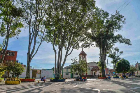 A quiet neighborhood square, surrounded by tall trees that filter the sunlight and cast shadows on the ground. In the center stands a small church with a white facade and a red-tiled bell tower, lending a traditional air to the surroundings.