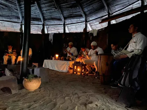 Un grupo de personas sentadas alrededor de un fuego en una cabaña, estilo maloka, compartiendo historias y disfrutando del calor.