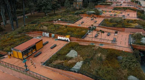 Aerial view of an urban park with wide, reddish-toned pathways surrounded by gardens and diverse vegetation. Benches, railings, and stepped terraces are visible, along with a small kiosk and a sign that reads ""Bogotá."" The surroundings are green and well-organized, with areas for rest and walking.
