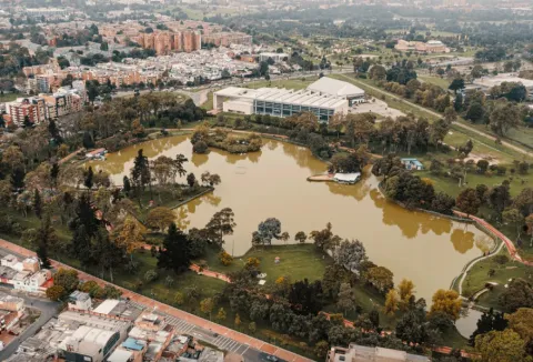 Aerial view of an urban park with a large lake surrounded by trees, pedestrian paths, and green spaces, with city buildings and neighborhoods in the background.