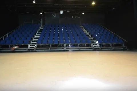 Interior of a theater with metal bleachers and rows of blue chairs arranged in tiers. The stage is visible in the foreground, empty and well-lit, while the background is dark, with technical lighting on the ceiling. The space conveys an atmosphere prepared for theatrical performances.