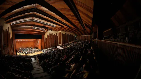 Vista frontal panorámica del interior de un gran auditorio durante un concierto sinfónico. En el escenario de madera se encuentra una orquesta con numerosos músicos e instrumentos, mientras el público llena las graderías y platea a ambos lados. El recinto destaca por su techo curvo de madera con vigas visibles y una iluminación cálida que enmarca el escenario.