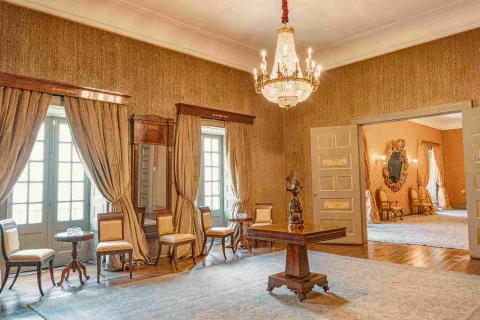 Interior de un salón elegante de estilo clásico, con paredes decoradas en tonos dorados y un candelabro de cristal colgando del techo. Hay muebles de madera y sillas tapizadas junto a ventanas con cortinas largas, y en el centro una mesa con una escultura. A través de una puerta abierta se observa otra sala con decoración similar y un gran espejo ornamental, creando un ambiente histórico y sofisticado.
