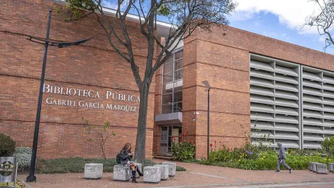 Fachada de ladrillo de la Biblioteca Pública Gabriel García Márquez, con su nombre visible en letras grandes. Frente al edificio hay un sendero peatonal rodeado de jardines y árboles; una mujer está sentada en un bloque de concreto mirando su celular y otra persona camina cerca.