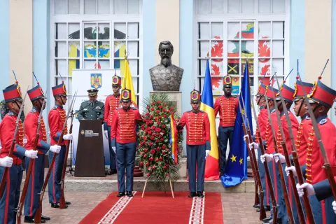 Military ceremony with a uniformed honor guard in front of a commemorative bust, accompanied by flags and a floral offering in the courtyard of a historic building.