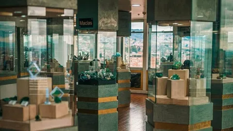 A museum exhibition hall with several glass display cases exhibiting emeralds and minerals on pedestals. The pieces are arranged in columns within a well-lit space, and in the background, a window offers a view of the city.