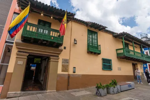 Fachada de una casa colonial de dos pisos con paredes color crema, balcones y ventanas de madera pintadas de verde. Sobre la entrada cuelgan las banderas de Colombia y de Bogotá. El edificio tiene techo de tejas de barro y una placa conmemorativa en la pared, mientras algunas personas caminan por la acera frente al lugar.