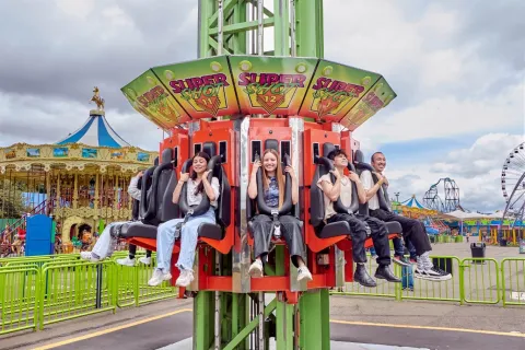 A group of young people sit on a mechanical ride at an amusement park, secured with harnesses while awaiting the vertical launch. In the background, other attractions such as a carousel and a Ferris wheel can be seen.