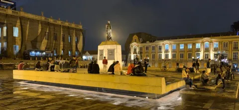 A public square at night with an illuminated statue in the center, surrounded by people sitting and conversing on a platform. The surrounding historic buildings are illuminated, highlighting their colonial architecture. The damp ground reflects the warm lights, creating a lively and inviting urban atmosphere at night.