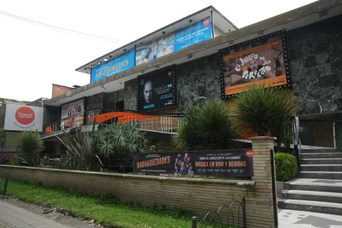 Facade of a cultural building or theater in an urban area, with advertising posters for plays at the top. The building has two levels, brick and stone walls, and an entrance with a black metal gate. A small garden with ornamental plants and a tree-lined walkway are visible in front, to the right.