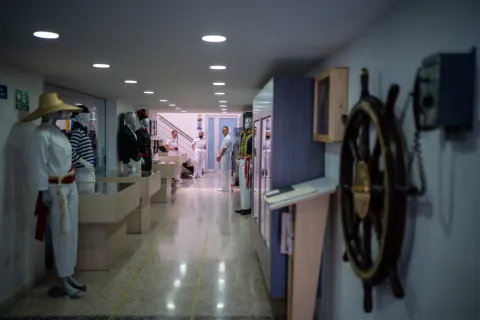 Pasillo interior de un museo o espacio expositivo con temática naval. A ambos lados se observan vitrinas y maniquíes vestidos con uniformes marítimos tradicionales, mientras al fondo varias personas con uniforme blanco conversan cerca de una escalera. En primer plano, a la derecha, destaca un timón de barco colgado en la pared, y la iluminación es moderna y uniforme, creando un ambiente ordenado y educativo.