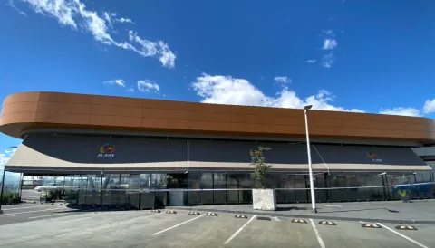 La imagen muestra un moderno edificio comercial de diseño contemporáneo, caracterizado por sus líneas curvas y una fachada elegante en tonos cálidos, que contrastan con el cielo azul intenso salpicado de nubes blancas.
