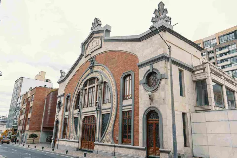 La imagen muestra la fachada de un edificio histórico de estilo ornamental, con detalles en ladrillo y piedra que destacan en el entorno urbano. Su diseño elegante contrasta con construcciones modernas cercanas, mientras algunas personas caminan por la calle, aportando vida a la escena bajo un cielo nublado.