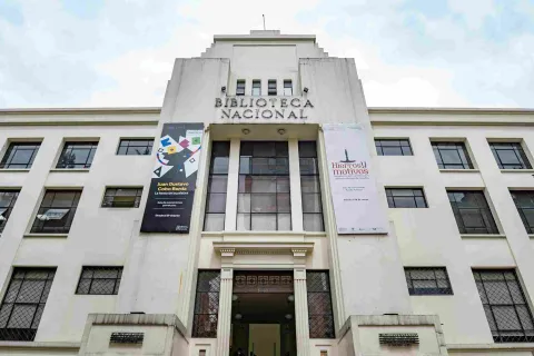 La imagen muestra la fachada de un edificio institucional de estilo sobrio y simétrico, identificado como la Biblioteca Nacional. Su diseño en tonos claros y líneas verticales resalta la entrada principal, flanqueada por grandes ventanales y banners informativos.