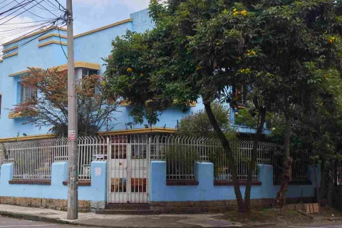 La imagen muestra una casa de dos pisos pintada en tonos azul claro con detalles amarillos, rodeada por una reja blanca. Al frente hay árboles que aportan sombra y vegetación, mientras un poste y cables eléctricos evidencian el entorno urbano. La escena transmite tranquilidad en un barrio residencial.