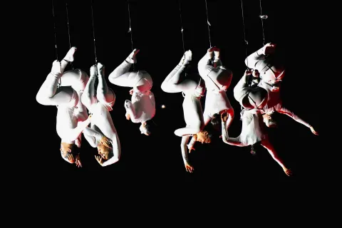 Grupo de acróbatas vestidos de blanco suspendidos boca abajo con arneses, alineados en el aire sobre un fondo negro y con iluminación dramática que resalta sus cuerpos y poses.