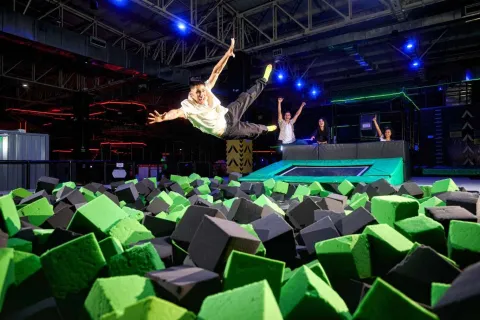 People jump and play in a foam pit, while one of them performs a somersault in an indoor trampoline park.
