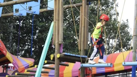 A child wearing a helmet and harness crosses a rope bridge in an outdoor adventure course, surrounded by colorful play structures.
