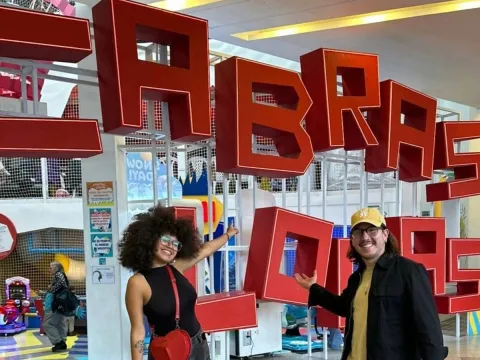 Two people smile and pose in front of large red letters spelling "CABRAS LOCAS" in an indoor play area.