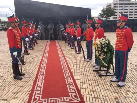 Guardia de honor con uniformes rojos custodia un monumento, junto a una ofrenda floral y una alfombra roja.
