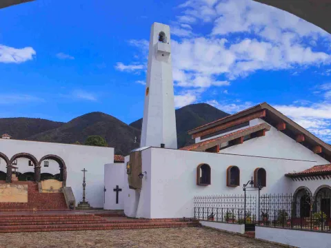 Iglesia de arquitectura blanca con campanario alto, rodeada de montañas y cielo azul, con una plaza de piedra en primer plano.