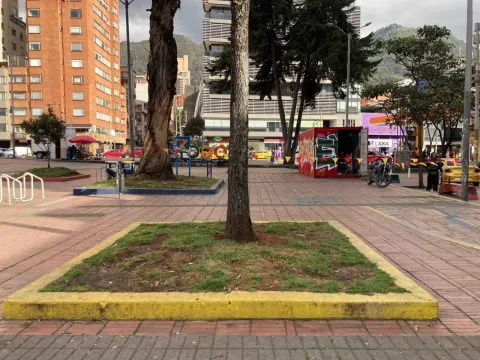 An urban plaza surrounded by buildings and trees in Bogotá, with pedestrian areas, mountains in the background, and an everyday city atmosphere.