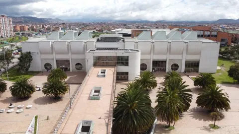 Vista exterior de una biblioteca pública de gran tamaño, con arquitectura moderna, rodeada de palmeras y una plaza amplia, con la ciudad de Bogotá extendiéndose al fondo bajo un cielo nublado.