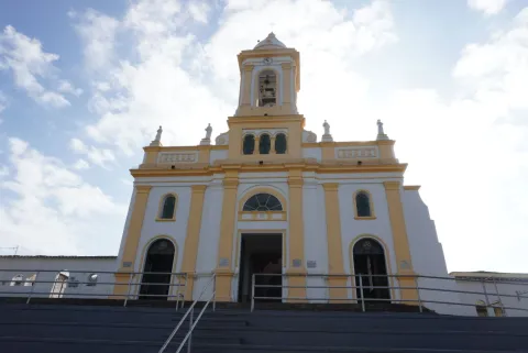 Facade of a colonial-style church in white and yellow tones, with a central tower and steps, seen from below against a partly cloudy sky.