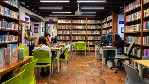 Sala de biblioteca con estanterías llenas de libros y mesas de estudio, donde varias personas leen y trabajan en un ambiente tranquilo y organizado.