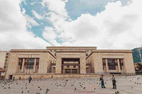 Fachada de estructura con tres edificaciones en piedra. Hay un policia conversando con un hombre y varias palomas en el suelo.