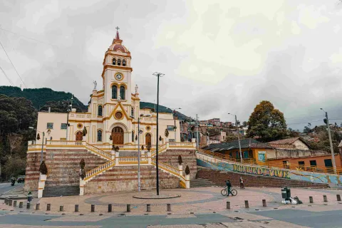 A wide shot of the facade of a light brown church, with mountains in the background. In the foreground, there is a person riding a bicycle.