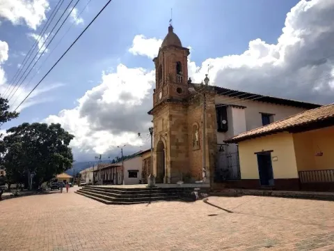 Town church with a stone tower and bell tower, located next to a large plaza, with colonial houses and a partly cloudy sky.