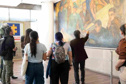 Un grupo de visitantes observa un gran mural mientras un guía señala detalles de la obra durante un recorrido en un espacio museístico.