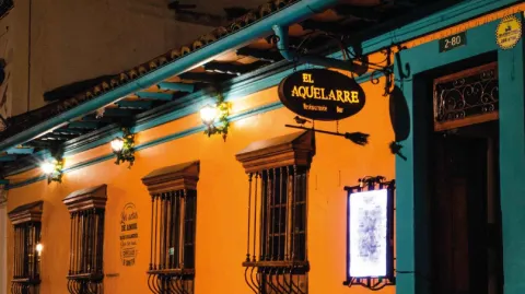 Fachada nocturna de una casa colonial de paredes amarillas, marcos azules y letrero luminoso de El Aquelarre Bar.