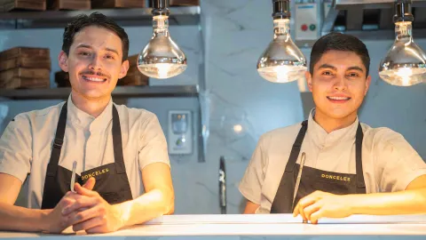 Dos jóvenes chefs sonrientes con delantal negro de Donceles posando tras el mostrador de iluminación cálida.