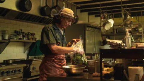 Un chef con una gorra de béisbol y un delantal de cocina prepara verduras en una bolsa de plástico en una cocina rústica.