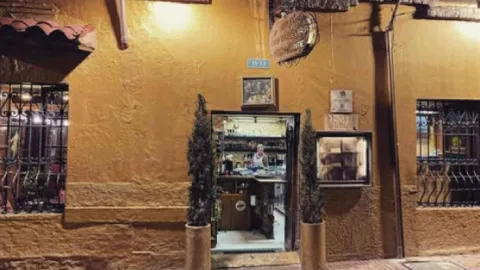 Exterior de casa colonial de paredes amarillas en la noche con plantas a la entrada y letrero del establecimiento.