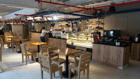 Mostrador de cafetería con vitrina exhibidora de postres, barista preparando café y mesas de madera alrededor. 