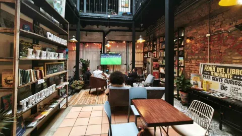 Patio interior con zona de librería, estantes con libros, mesas de madera y clientes viendo una televisión