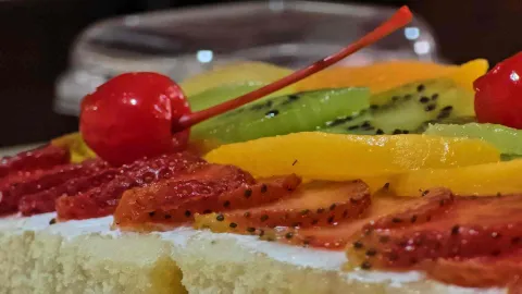 Primer plano de postre cubierto con rodajas de fresa, kiwi, melocotón y una cereza sobre bizcocho blanco.