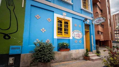 FOTO 1: Edificio con fachada azul, ventana y puerta amarillas. El letrero circular del restaurante y plantas decoran la entrada.
