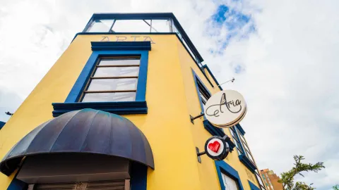 Foto 1: Fachada de un edificio amarillo de esquina con marcos de ventanas azules, marquesina oscura y letreros circulares de la marca Aria.