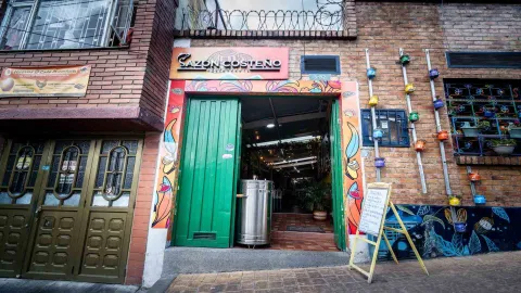 Foto 2: Fachada del restaurante Sazón Costeño con puerta verde abierta, barril ahumador y macetas coloridas en la pared.