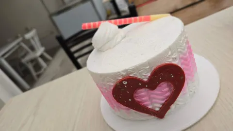 Pastel cilíndrico decorado con crema blanca y rosada, detalle de corazón de chocolate rojo y espiral dulce. 