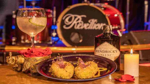 FOTO PORTADA REVELLION: Un cóctel de ginebra y Hendrick's, junto a dos aperitivos empanizados, una vela y pétalos de rosa, con una batería de fondo que dice "Rebellion".