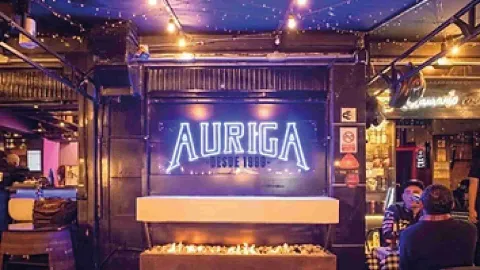 Foto 1: Letrero luminoso de "AURIGA DESDE 1998" sobre una chimenea encendida en el interior del bar. 